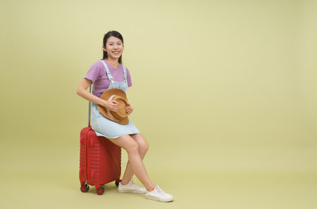A young Asian woman smiles while seated on a bright red suitcase, holding a straw hat. She wears a casual outfit, embodying a sense of excitement for upcoming travel.の写真素材