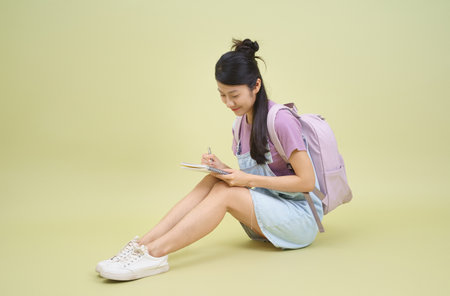 A girl sits on the floor, wearing a light blue dress and a pastel backpack. She enjoys writing in her notebook, showcasing a focused expression and relaxed pose.の写真素材