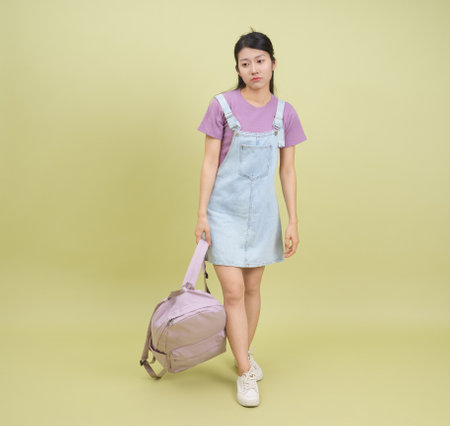 A young woman stands against a pale yellow background, wearing a light denim dress over a purple t-shirt. She carries a light purple backpack and appears contemplative and relaxed.の写真素材