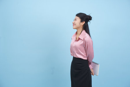 A young woman wearing a pink blouse and black skirt stands with a slight smile, holding a notebook behind her back. The bright blue background adds a cheerful atmosphere to the scene.の写真素材