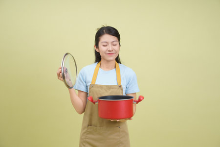 A young Asian woman stands in a cheerful kitchen, holding a red pot with a lid and smiling contentedly. Her apron hints at recent cooking activity and enjoyment in the moment.の写真素材