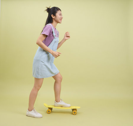 A girl skates joyfully on a yellow skateboard while dressed in a denim dress and white shoes. Her casual outfit and cheerful expression highlight a fun, carefree moment.の写真素材