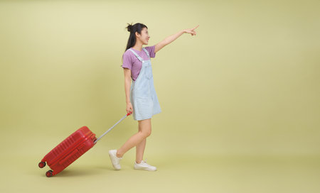 A young Asian woman in casual attire is happily pulling a bright red suitcase. She points enthusiastically, suggesting excitement for adventures ahead in a simple studio backdrop.の写真素材