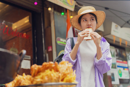 A young woman in a straw hat bites into a tasty snack while standing in a vibrant street market. Colorful signs and lights create a lively backdrop for her culinary experience.の写真素材