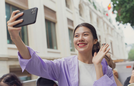 A cheerful young woman with long hair smiles and waves while capturing a selfie with her smartphone. She is surrounded by friends near a historic building, enjoying the moment.の写真素材