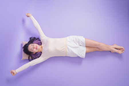 A young woman is lying comfortably on a purple surface, arms extended outward and eyes closed. She appears to be embracing a moment of relaxation, savoring stillness in a peaceful indoor environment.の写真素材