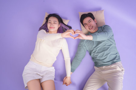 A young couple lies on a purple mat, holding hands and forming a heart shape with their fingers. Their playful expressions reflect happiness and affection in a lighthearted moment.の写真素材