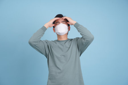 A young Asian man stands against a light blue wall, wearing a protective mask while holding his head in a gesture of stress and frustration, conveying a sense of exhaustion and anxiety.の写真素材