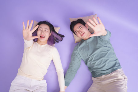 A young couple lies side by side on a cozy surface, their hands intertwined. They are both smiling, enjoying a playful moment together amidst a soft purple backdrop.の写真素材