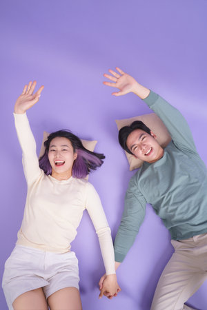 Two young individuals, a man and a woman, are lying side by side on a purple surface with pillows, smiling broadly while holding hands and waving their arms joyfully.の写真素材