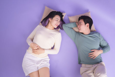 A young Asian couple lies on a purple surface, using pillows for support. They smile and engage in an intimate, light-hearted conversation while enjoying their time together.の写真素材