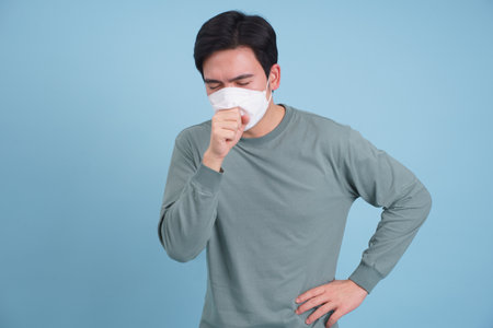 A young man in a gray long-sleeve shirt coughs while wearing a mask, showing discomfort. He stands...