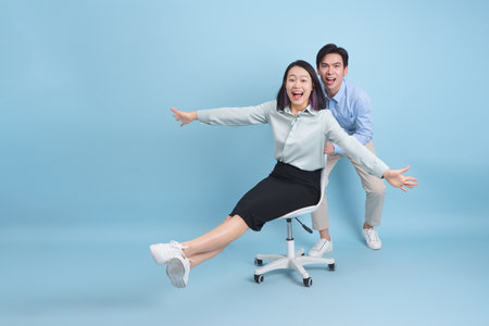 Two Asian coworkers share a joyful moment in a bright blue background, with one playfully pushing the other in a swivel chair while laughing and having fun together.の写真素材