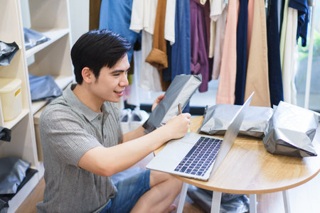 A young Asian man sits at a wooden table, focusing on his laptop while preparing packages in a casual setting surrounded by clothing. He writes on packaging materials with a pen.の写真素材