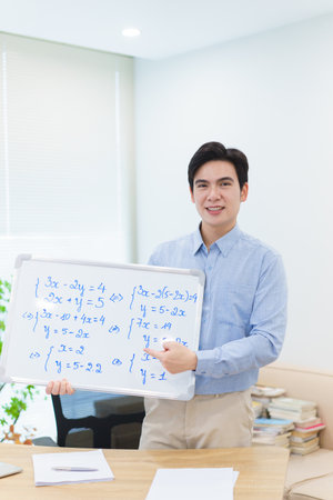 A young Asian math tutor stands confidently in a well-lit classroom, pointing to a whiteboard filled...