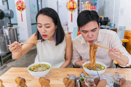 A man and woman are immersed in enjoying bowls of steaming noodle soup at a laid-back Asian eatery,...