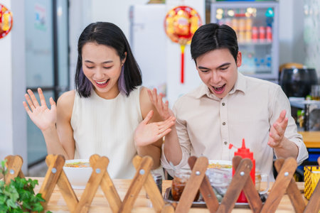 Joyful couple shares a moment of excitement as they savor a festive meal. They are seated at a table adorned with symbolic decorations, showcasing a blend of modern and traditional elements.の写真素材