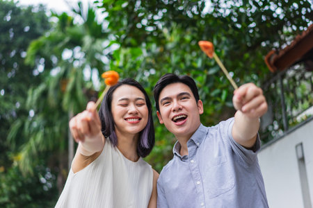 A young couple is smiling and playfully holding food skewers, surrounded by palm trees and greenery. They appear happy, enjoying each other's company on a warm day.の写真素材