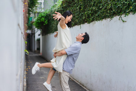 A happy couple shares a playful embrace in a narrow alley, with one partner lifting the other as they laugh together. Their surroundings are filled with greenery, creating a vibrant atmosphere.の写真素材