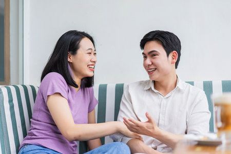 A young man and woman are sharing laughter and an animated discussion while sitting comfortably in a modern cafe. The bright, welcoming atmosphere enhances their connection.の写真素材