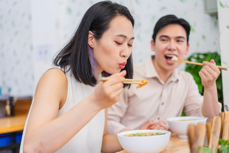 A woman delicately sips her noodles while a man looks on, both engaged in a savory meal at a vibrant...