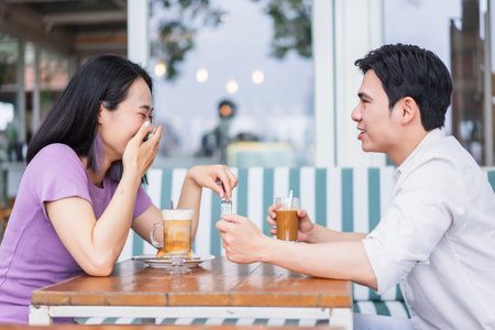 A young couple shares a moment of joy over drinks at a cafe. They engage in playful conversation, surrounded by a lively atmosphere, enjoying the warm sunlight.の写真素材