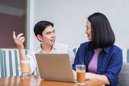 A young couple is engaged in a lively conversation while working on a laptop at a cafe. They share smiles and drinks, enjoying the relaxed atmosphere and each other's company.の写真素材