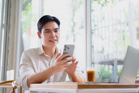 A young man wearing a casual shirt smiles while interacting with his smartphone at a cafe. A...