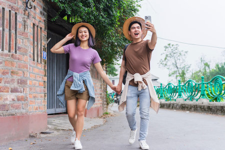 Young couple walks hand in hand down a picturesque street, smiling and taking a selfie. They are dressed casually for a delightful day exploring their surroundings together.の写真素材