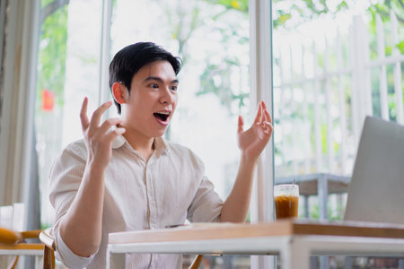 A young Asian man sits at a table in a bright cafe, engaging in an animated video call. He shows excitement, with hands raised and a surprised expression, while enjoying a cold drink.の写真素材