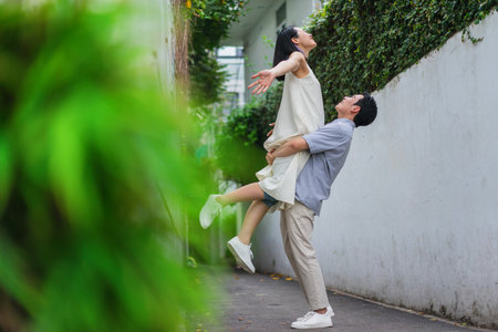 Two people share a happy moment in a vibrant alley surrounded by greenery. The man lifts the woman affectionately as they enjoy a warm sunny day in the city, celebrating their love.の写真素材