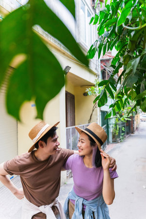 A couple strolls together in a charming neighborhood, wearing straw hats and casual clothing. They share playful laughter while enjoying each other's company amidst lush greenery.の写真素材