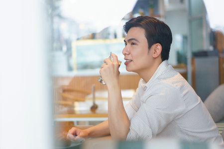 A young Asian man sits comfortably at a stylish cafe, sipping a beverage while admiring the lively atmosphere around him during a bright afternoon.の写真素材