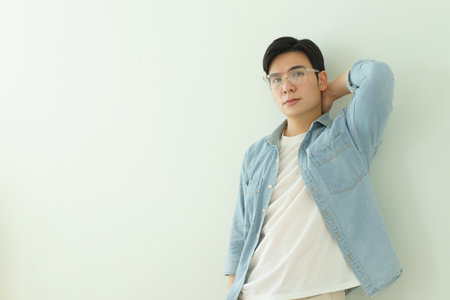 A young Asian man stands confidently against a soft light wall. He wears a denim shirt over a white t-shirt, presenting a modern and stylish look. The atmosphere is bright and relaxed.の写真素材