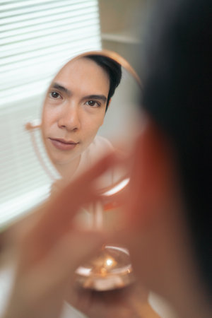 A young Asian man is focused on his reflection while applying skincare in a bright room. Sunlight filters through the blinds, highlighting his careful grooming process.の写真素材