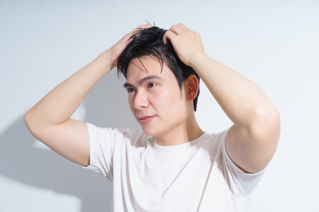 A young Asian man is seen styling his hair with his hands while standing against a plain backdrop. He appears focused and relaxed, dressed casually in a simple white T-shirt.の写真素材