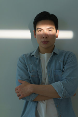 A young Asian man stands confidently with his arms crossed, illuminated by a narrow beam of natural light. The minimalist background emphasizes his calm expression and style.の写真素材
