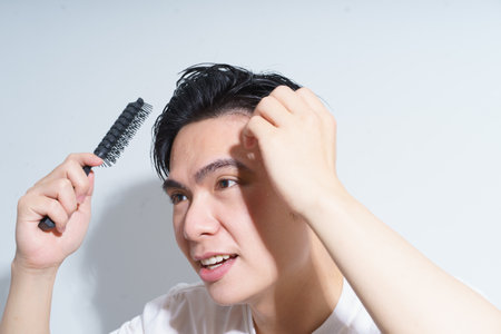A young Asian man is brushing his hair with a black comb, smiling while looking in the mirror. The simple background emphasizes his focused grooming routine in a well-lit room.の写真素材