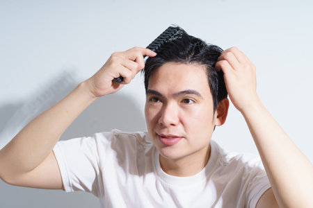 A young Asian man is seen styling his hair with a comb, focusing intently in front of a bright white background. Natural light highlights his features and clean attire.の写真素材