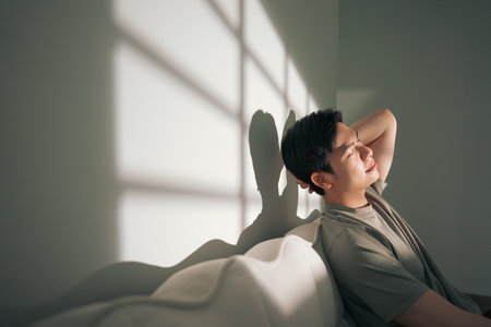 A young Asian man leans back against a soft couch, soaking in the warm afternoon light filtering through the window. Shadows create a calming atmosphere as he relaxes.の写真素材