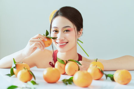 A cheerful young woman holds a small orange while sitting among a bountiful arrangement of fresh oranges. The vibrant colors and natural lighting create a joyful atmosphere.の写真素材