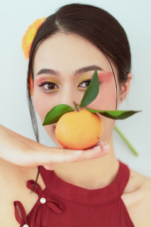 In a vibrant, sunny setting, a young woman balances an orange on her palm while playfully posing. Her makeup features bright colors that complement the lively atmosphere.の写真素材
