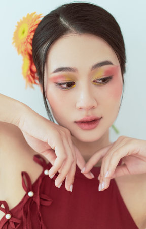 A young woman of Asian descent displays stunning makeup with colorful eyeshadow and a flower accent in her hair. Her gentle expression complements the warm, inviting atmosphere.の写真素材