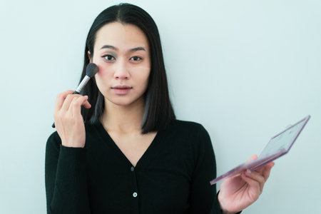 A woman stands in a minimalist environment, applying blush to her cheeks using a brush. She appears focused, with a calm expression, dressed in a black top.の写真素材