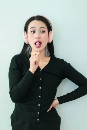 A girl with dark hair and light highlights stands against a plain wall, wearing a black sweater and pink headphones. She playfully holds a lollipop to her lips, lost in the music.の写真素材