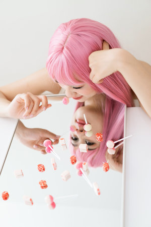 A young woman with vibrant pink hair leans over a mirror, playfully reaching for colorful candies scattered on its surface, showing a moment of joy and creativity in a bright indoor space.の写真素材