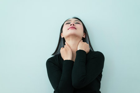 A young woman with straight hair is sitting against a light wall, holding her neck. She appears to be lost in thought, expressing deep emotion and introspection in her pose.の写真素材