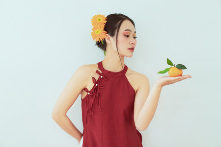 A woman with flowers in her hair gracefully poses while holding a mandarin orange in her palm. The...
