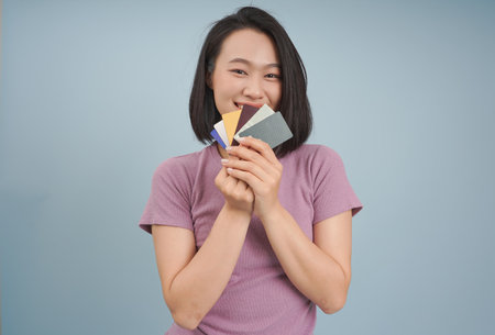 A young woman with straight black hair and a cheerful expression holds a variety of colorful cards in her hands. She stands in front of a solid blue background, exuding joy and excitement.の写真素材