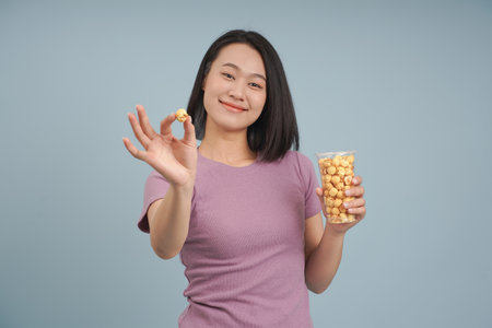 A cheerful young woman holds a piece of popcorn in one hand while offering a cup filled with popcorn...